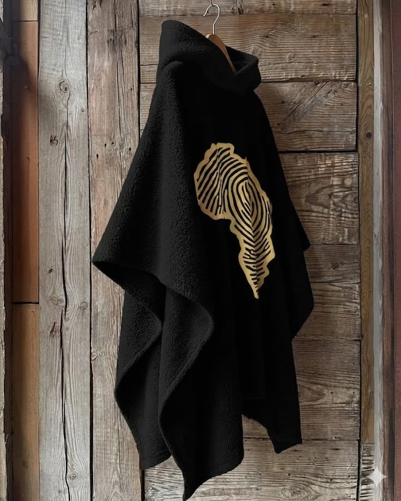 Black oversized hooded fleece cape featuring a large gold Africa-shaped fingerprint graphic on the back, hanging on a wooden hanger against a rustic wooden wall. Modern African-inspired poncho design with a relaxed silhouette and warm texture.